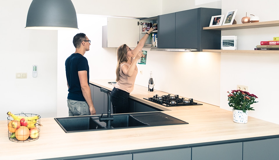 Un couple dans une cuisine moderne, l'un ouvrant un placard gris foncé, avec un plan de travail en bois clair et un panier de fruits.