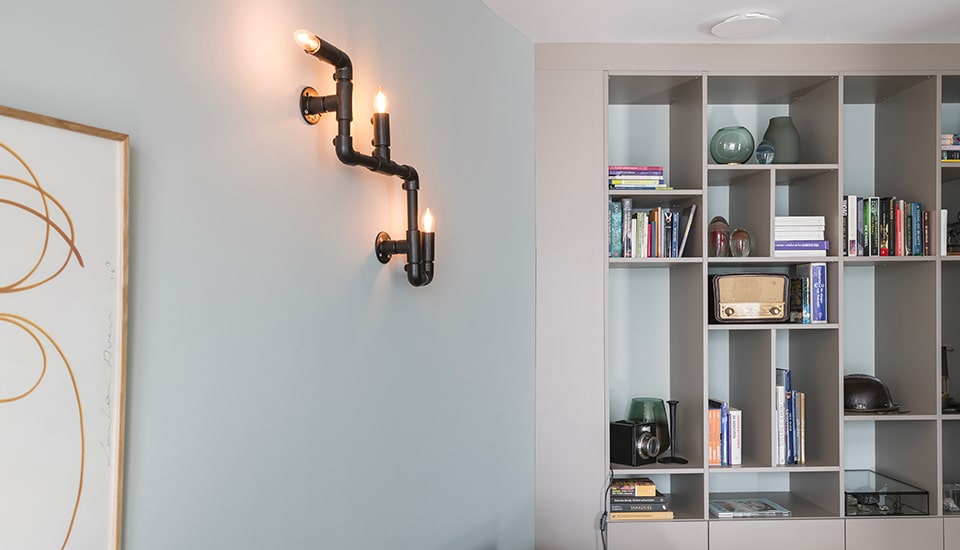 Une applique murale noire éclaire un mur gris à côté d'une bibliothèque contenant des livres, des vases et une radio vintage.