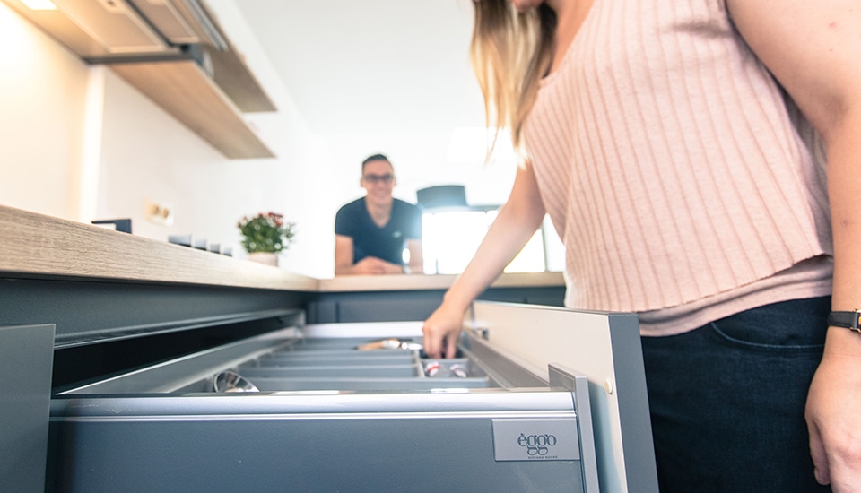 Une femme en haut rose ouvre un tiroir de cuisine gris, avec un homme flou à l'arrière-plan près d'un plan de travail en bois.
