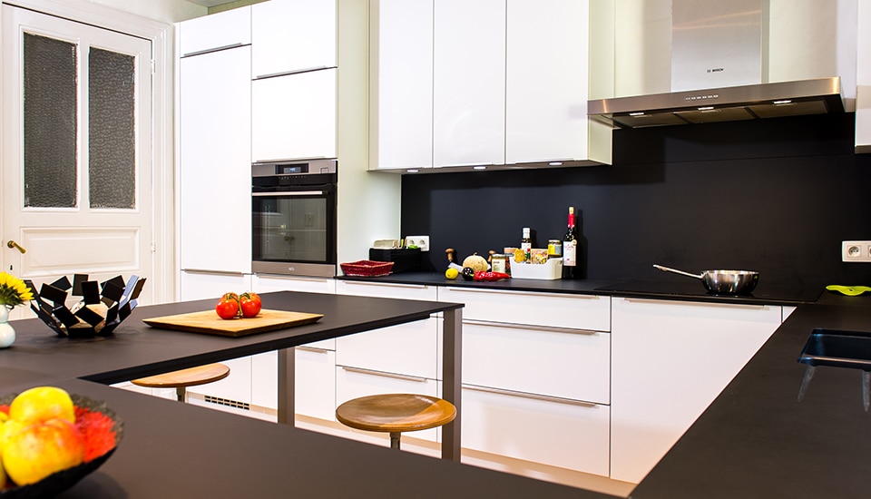 Cuisine moderne avec meubles blancs, plan de travail noir, tabourets en bois, et accessoires de cuisine, dont fruits et bouteilles, sur le comptoir.