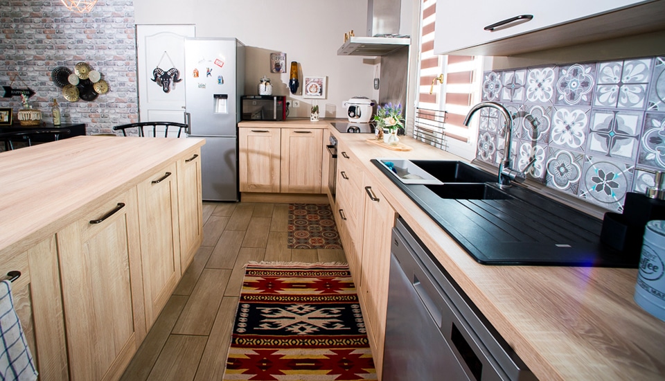 Cuisine moderne avec meubles en bois clair, plan de travail noir, carrelage à motifs et réfrigérateur argenté, éclairée par une fenêtre.