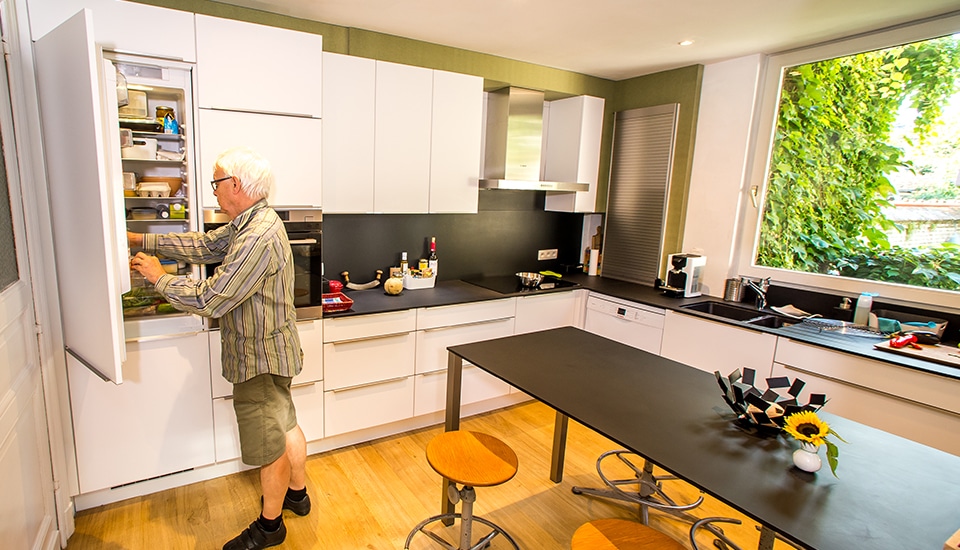 Un homme ouvre un réfrigérateur dans une cuisine moderne avec des placards blancs, un plan de travail noir et une grande fenêtre.