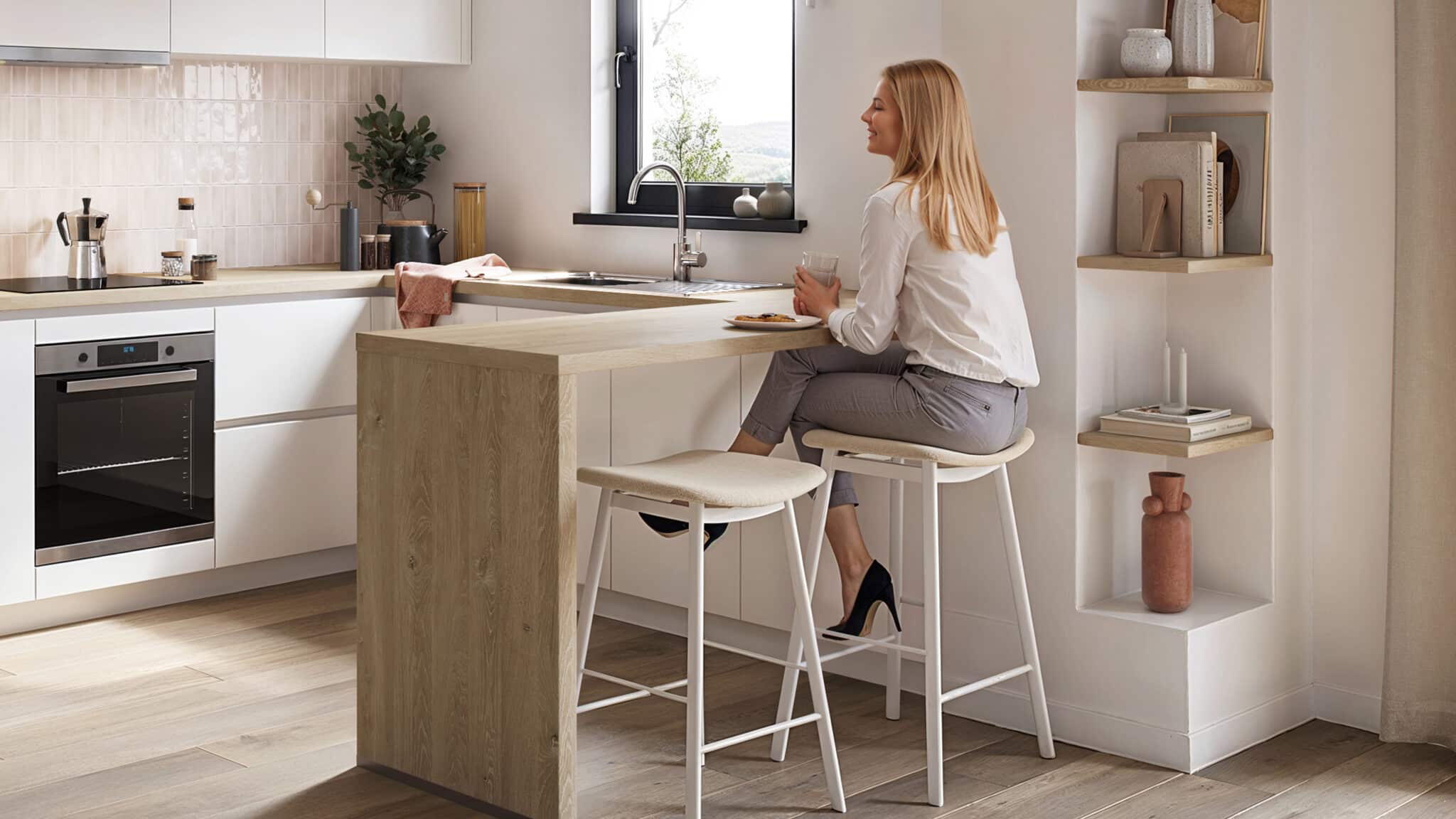 Une femme assise sur un tabouret de bar blanc dans une cuisine moderne avec plan de travail en bois clair et étagères décoratives.
