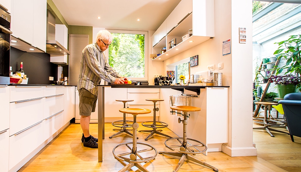 Un homme prépare des fruits dans une cuisine moderne avec des armoires blanches et des tabourets en bois devant un îlot central.