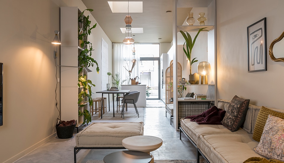 Salon moderne avec canapé beige, tables basses rondes et étagères blanches, plantes vertes et lampes suspendues, menant à une salle à manger.