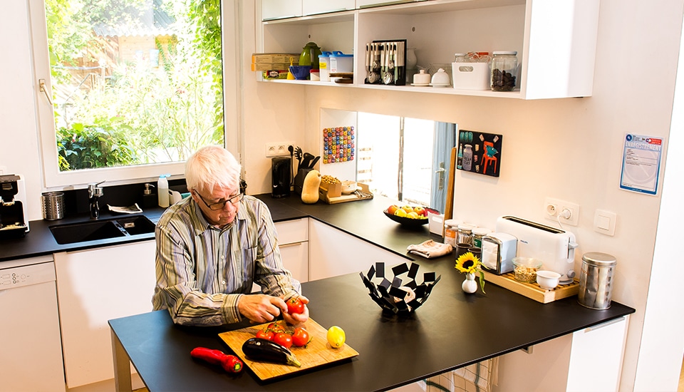 Un homme aux cheveux blancs prépare des légumes sur une planche à découper dans une cuisine moderne avec des étagères blanches.