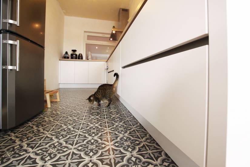 Cuisine moderne avec sol en carreaux à motifs gris, réfrigérateur en acier inoxydable et un chat tigré marchant près des armoires blanches.