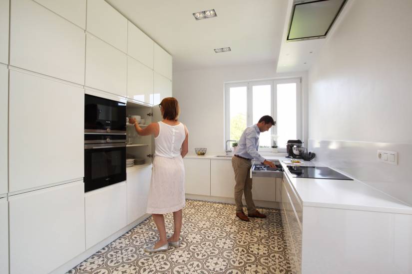 Deux personnes dans une cuisine moderne blanche avec un sol à motifs, l'une utilisant un four encastré, l'autre préparant à la cuisinière.