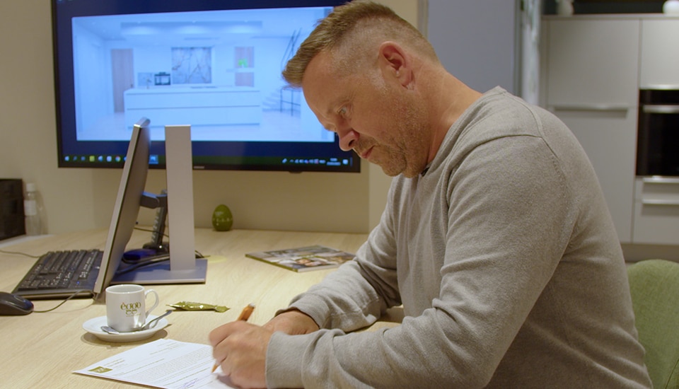 Un homme en pull gris écrit sur un document à un bureau, avec un écran affichant une cuisine moderne en arrière-plan.