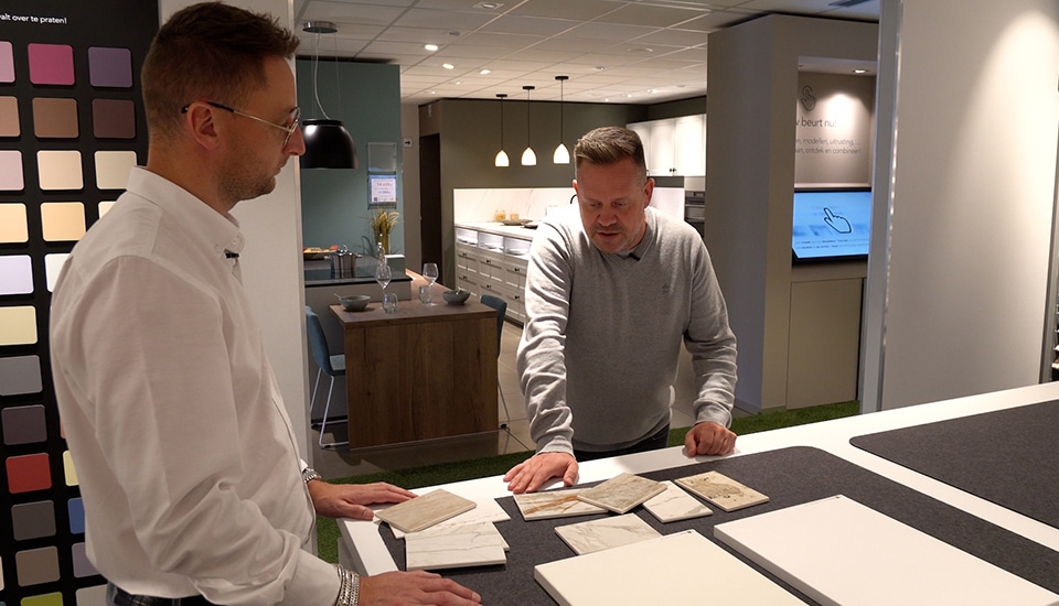 Deux hommes discutent devant un échantillon de matériaux de cuisine dans un showroom moderne avec un mur de couleurs et des luminaires suspendus.