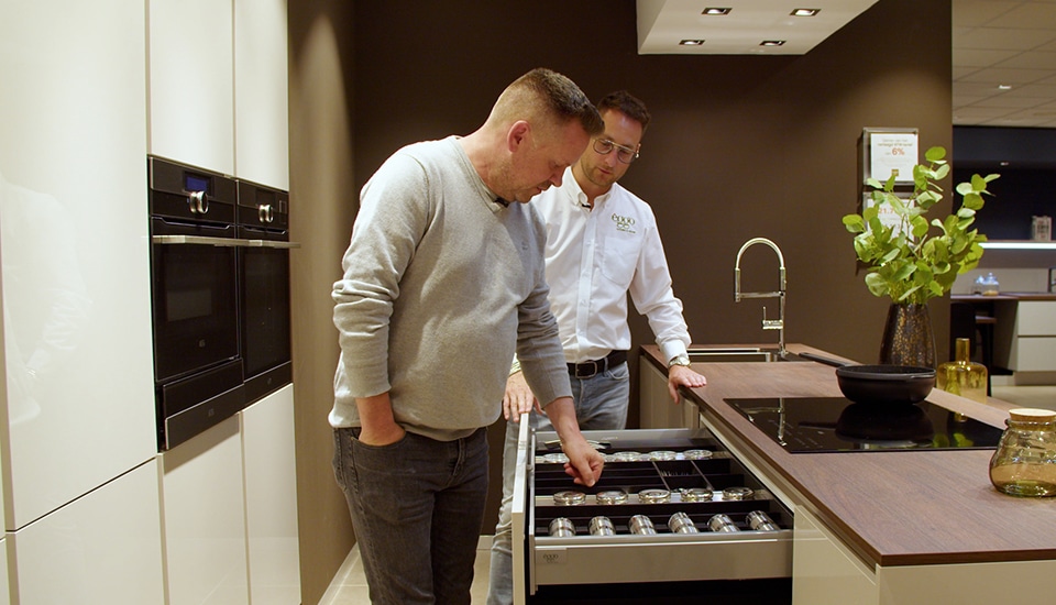 Deux hommes examinent un tiroir de cuisine rempli de bocaux, dans une cuisine moderne avec un plan de travail en bois et un évier en acier inoxydable.