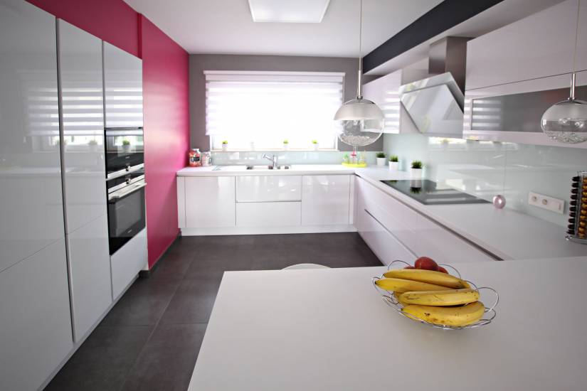 Cuisine moderne avec des armoires blanches, mur rose, et comptoirs épurés. Des bananes et une pomme sont posées sur une table blanche.