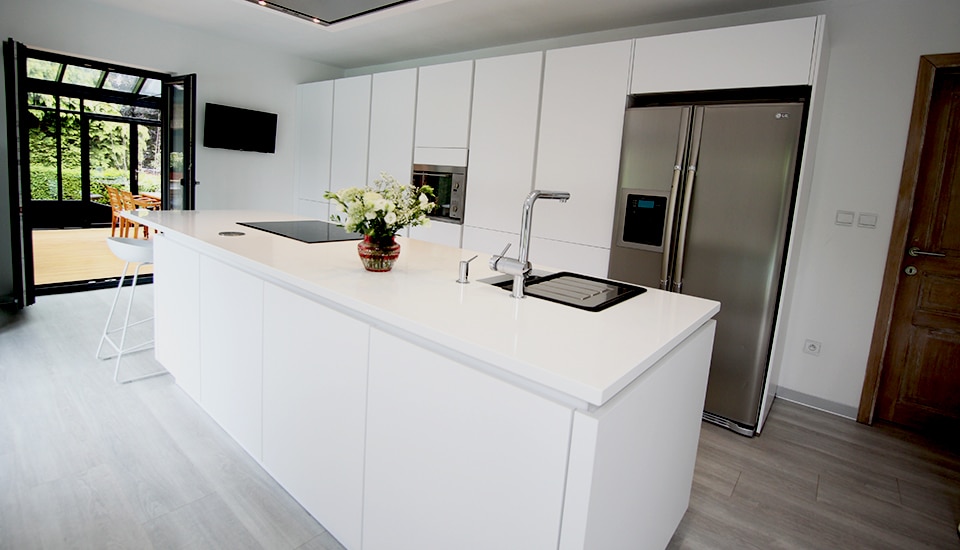 Cuisine moderne avec îlot central blanc, évier intégré, vase de fleurs blanches, et réfrigérateur en acier inoxydable à droite.