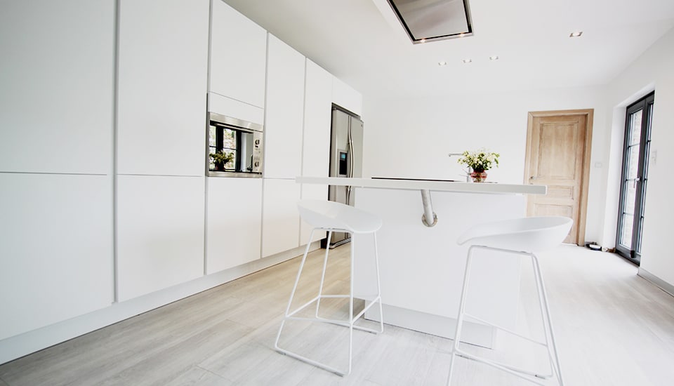 Cuisine moderne avec des armoires blanches, un îlot central et deux tabourets blancs, réfrigérateur en acier inoxydable et porte en bois clair.