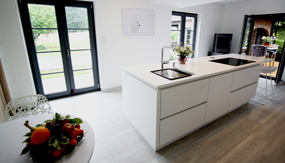 Cuisine moderne avec îlot central blanc, évier en inox, et plaque de cuisson noire. Fruits colorés sur table blanche et vue sur jardin.