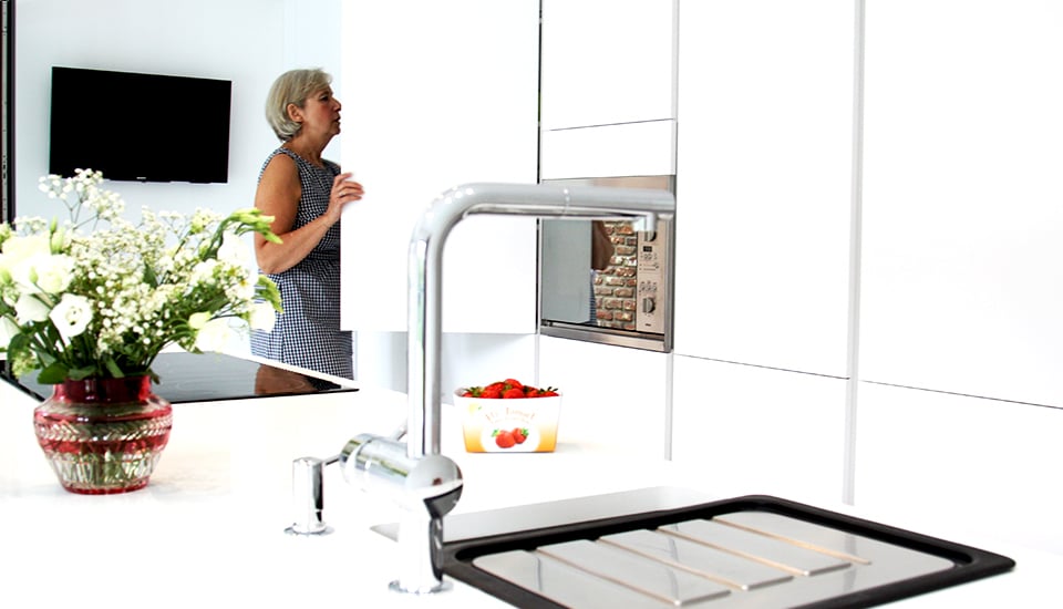 Un bouquet de fleurs blanches sur un plan de travail blanc, avec un évier en acier inoxydable et une femme ouvrant un placard.