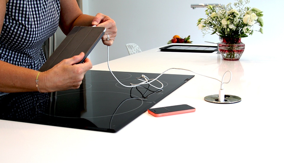 Une personne utilise une tablette connectée à une prise sur un plan de travail blanc, avec une plaque de cuisson noire et un vase de fleurs.