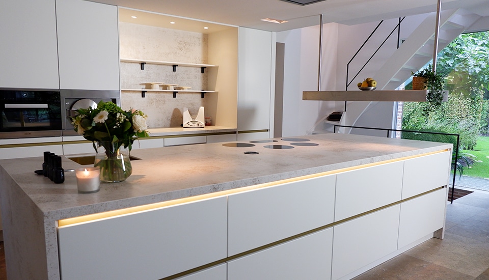 Îlot de cuisine moderne avec plan de travail en pierre, armoires blanches, vase de fleurs, bougie allumée et étagères avec vaisselle.