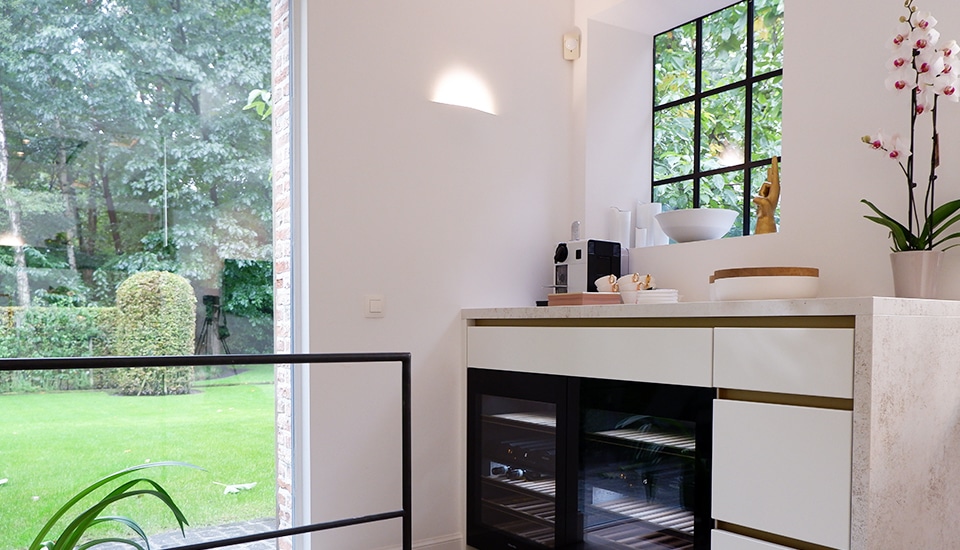 Cuisine moderne avec plan de travail beige, machine à café, vaisselle blanche, orchidée en pot et fenêtre donnant sur jardin verdoyant.