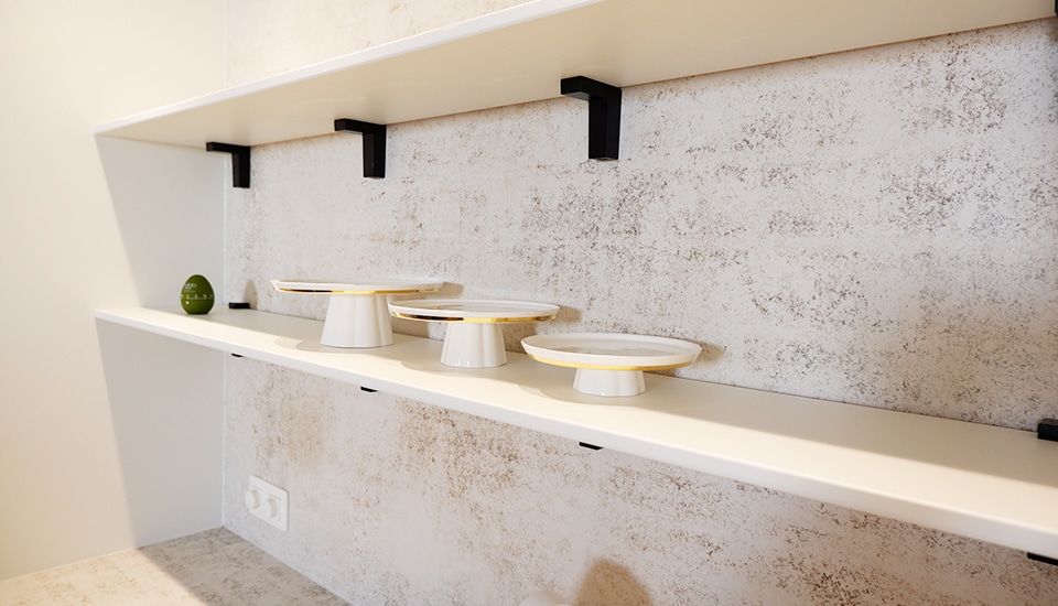 Étagères blanches supportant trois présentoirs à gâteaux à bord doré et un petit objet vert, fixées sur un mur texturé gris clair.