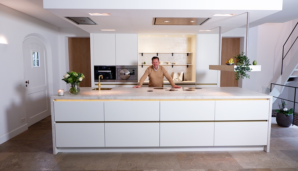 Cuisine moderne avec un îlot central blanc, des armoires intégrées, des étagères murales, et un homme souriant au centre.