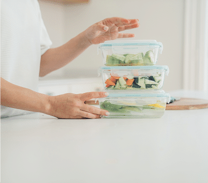 Des mains empilent trois boîtes transparentes contenant des légumes coupés sur un plan de travail blanc.