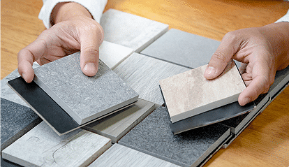 Des mains tiennent des échantillons de carrelage de différentes textures et couleurs sur une table en bois.