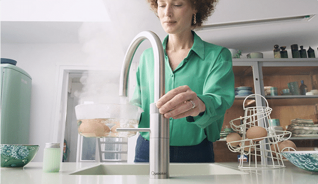 Une femme en chemise verte utilise un robinet moderne dans une cuisine, avec des œufs posés dans un support à côté.