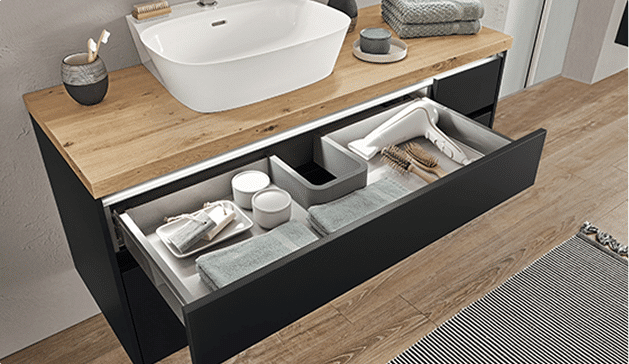 Tiroir de salle de bain noir ouvert sous un plan en bois avec lavabo blanc, contenant serviettes, brosses et accessoires de toilette organisés.