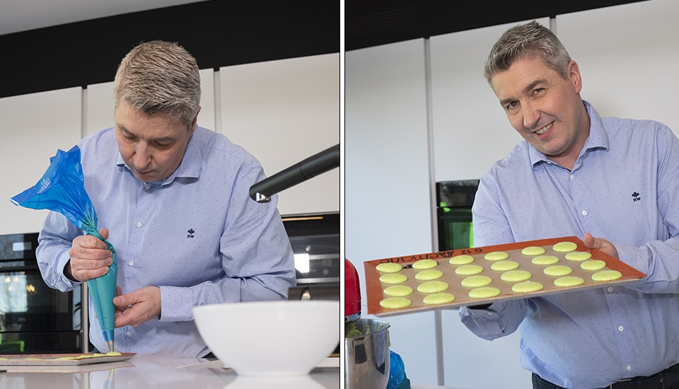 Un homme en chemise bleue prépare des macarons verts avec une poche à douille et présente un plateau garni dans une cuisine moderne.