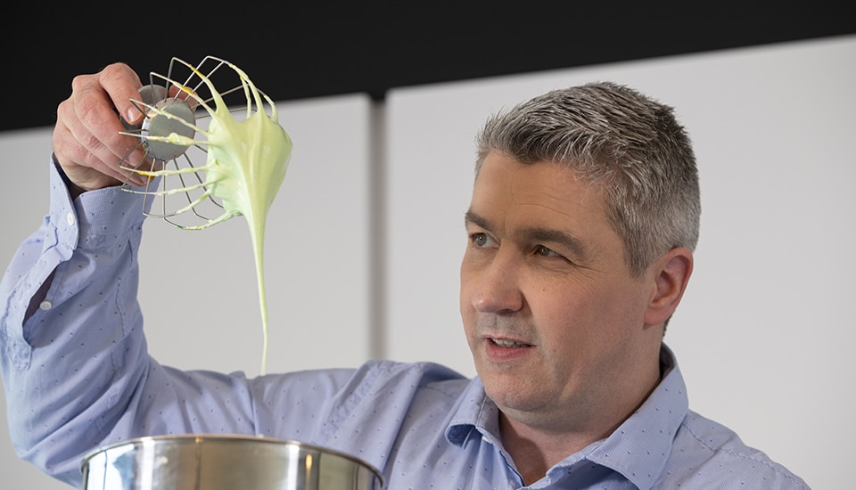 Un homme en chemise bleue tient un fouet avec une pâte verte au-dessus d'un bol en métal.