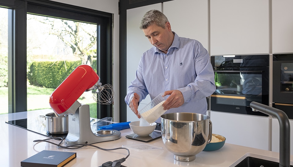 Un homme verse des ingrédients dans un bol blanc sur un plan de travail avec un robot de cuisine rouge et des appareils électroménagers en arrière-plan.