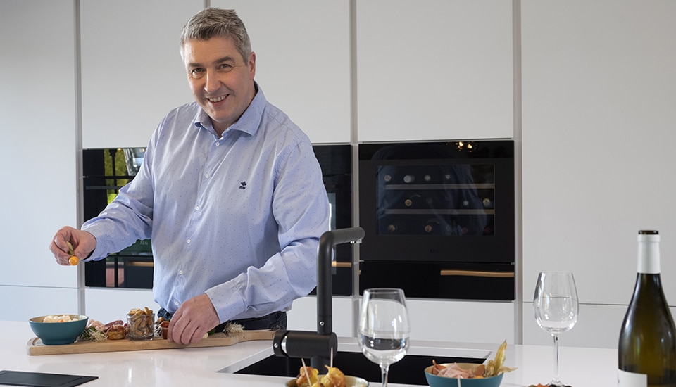 Un homme en chemise bleue prépare des amuse-bouches sur un plan de travail blanc, avec des verres à vin et une bouteille.