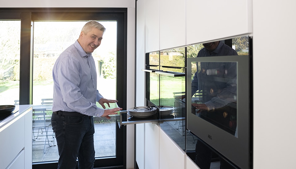 Un homme en chemise bleue utilise un tiroir de cuisine moderne avec des assiettes, près d'une grande fenêtre donnant sur un jardin.