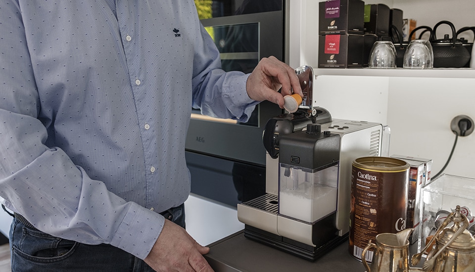 Un homme en chemise bleue insère une capsule dans une machine à café blanche sur un plan de travail, avec des boîtes de cacao à proximité.