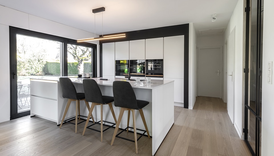 Cuisine moderne avec îlot central blanc, chaises noires, et grande baie vitrée donnant sur un jardin. Sol en bois clair et luminaires suspendus.