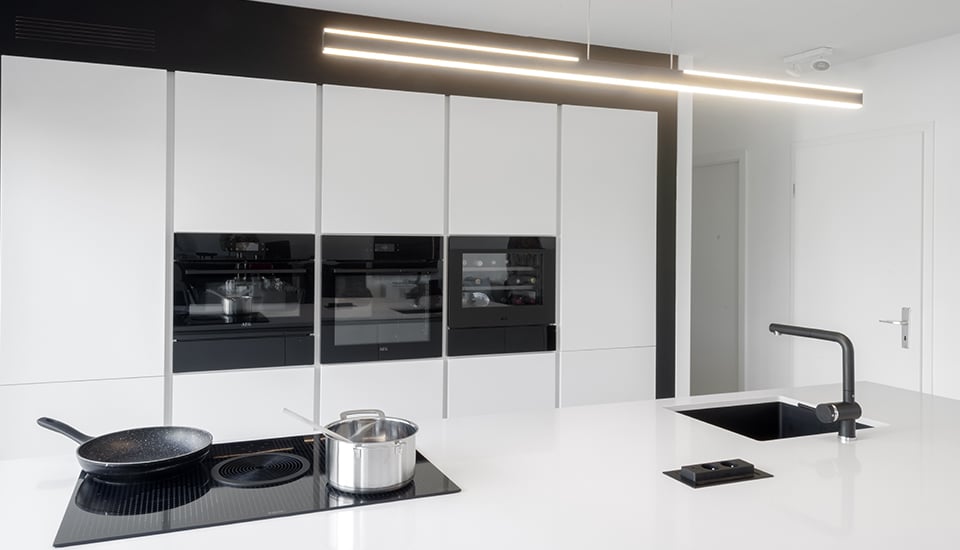Cuisine moderne avec placards blancs, plaque de cuisson noire, poêle, casserole en inox et évier noir sous un éclairage linéaire.