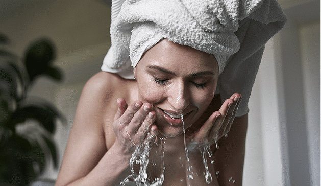 Une personne avec une serviette blanche sur la tête se lave le visage avec de l'eau, plante en arrière-plan.