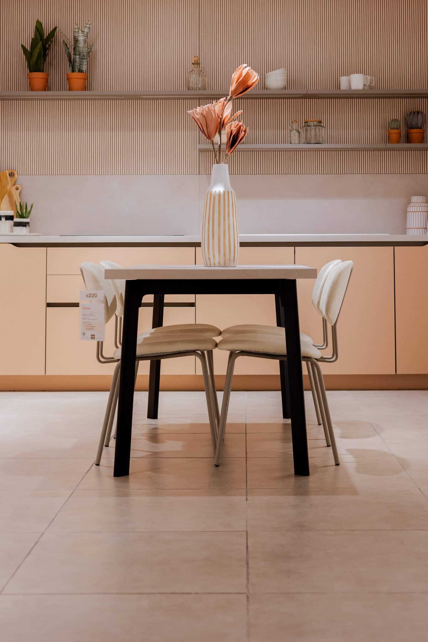 Une table avec un vase à fleurs roses est entourée de chaises beiges dans une cuisine moderne aux tons clairs.