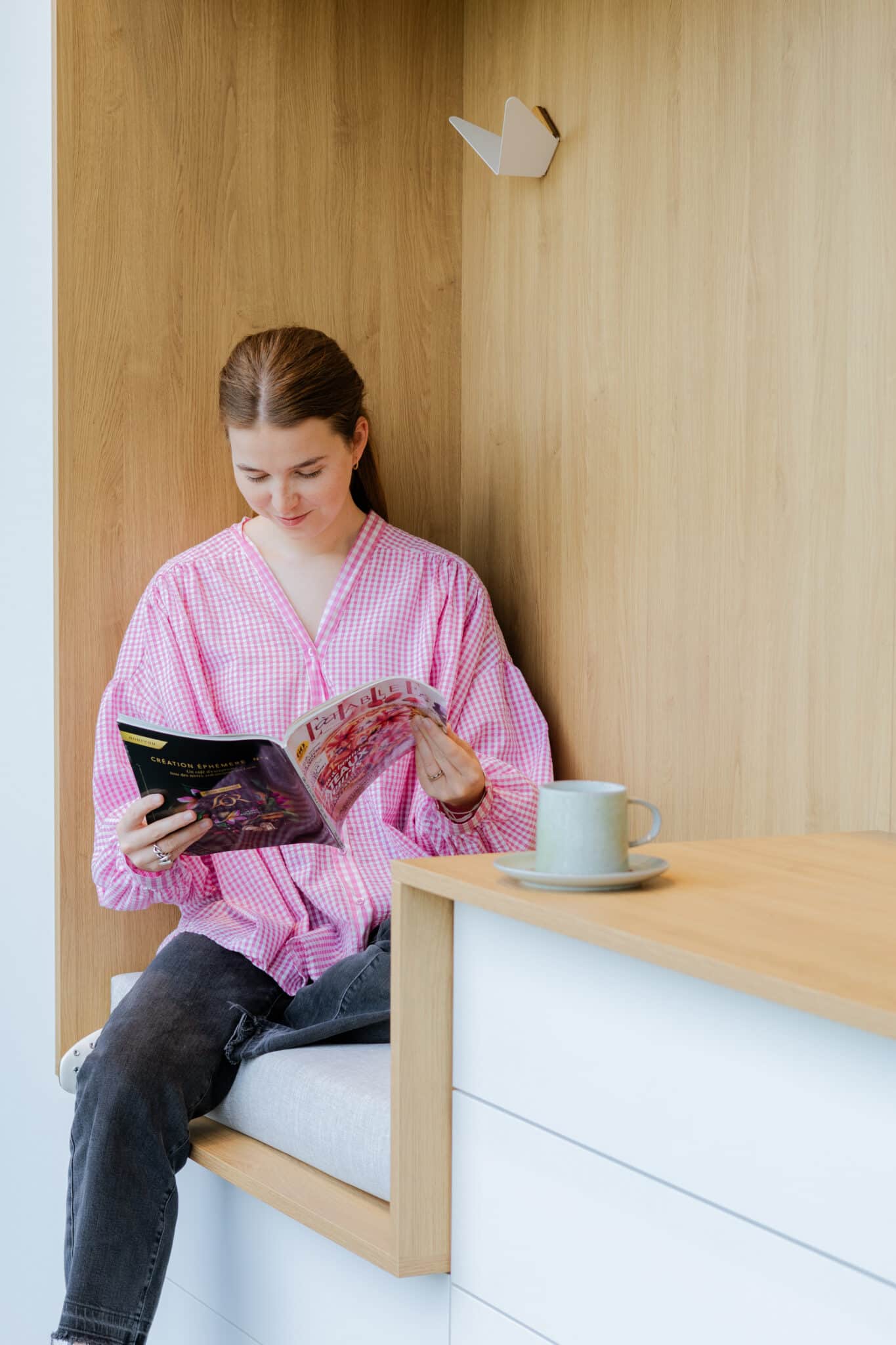 Une personne en chemise rose lit un magazine assise sur un banc en bois clair, avec une tasse verte posée à côté.