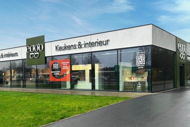 Magasin èggo avec façade en verre, affiches rouges et vertes, et intérieur visible. Bâtiment rectangulaire sur fond de ciel bleu.