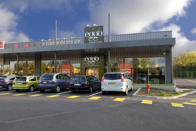 Façade d'un magasin èggo avec plusieurs voitures garées devant, affiches orange sur les vitrines, ciel bleu et nuages en arrière-plan.