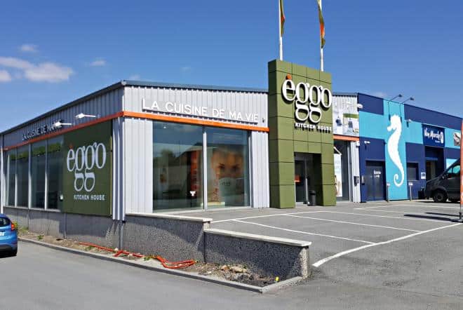 Magasin èggo avec façade grise et verte, logo visible, grandes vitrines et parking à l'avant sous un ciel bleu.