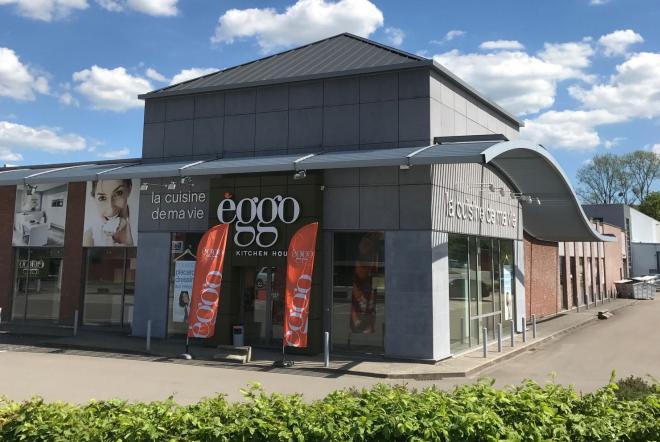 Façade d'un magasin èggo avec des murs en briques et panneaux gris, ornée de drapeaux orange, sous un ciel bleu.