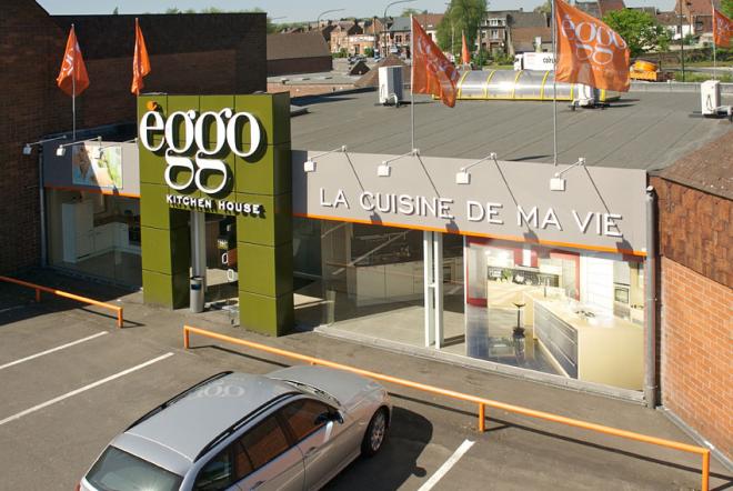 Façade d'un showroom èggo avec enseigne verte et slogan "La cuisine de ma vie", drapeaux orange, et voiture grise stationnée devant.