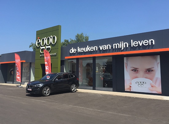 Un magasin Èggo avec une façade moderne, des drapeaux rouges, et une voiture noire garée devant, sous un ciel bleu clair.