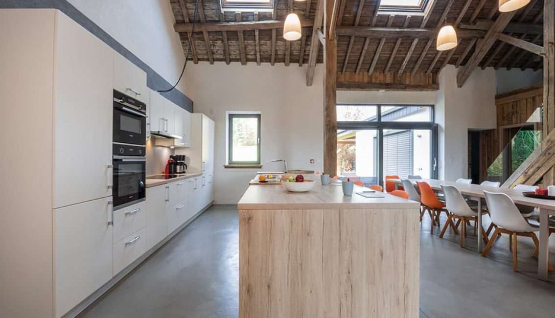 Cuisine moderne avec îlot central en bois clair, équipée de placards blancs, électroménager noir, et table à manger avec chaises orange et blanches.