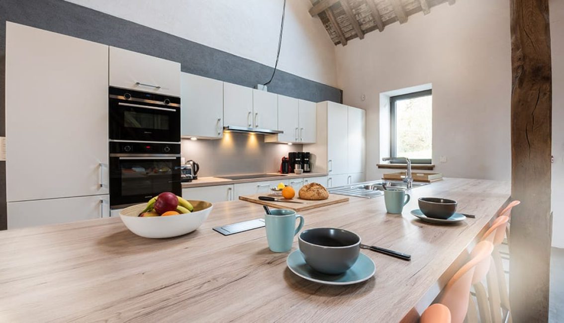 Cuisine moderne avec îlot central en bois clair, bols et tasses colorés, corbeille de fruits, et appareils encastrés dans des armoires blanches.