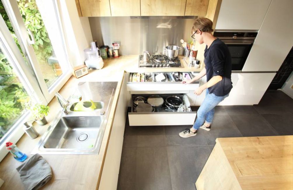 Une personne ouvre un tiroir de cuisine contenant des casseroles, à côté d'un évier en inox et d'une cuisinière à gaz.