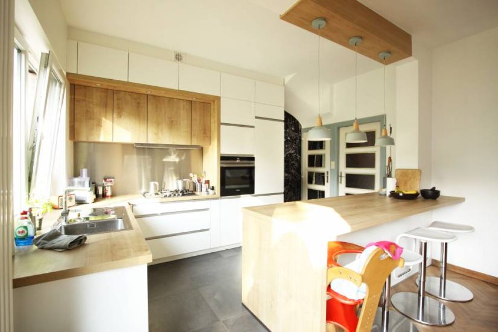 Cuisine moderne avec meubles en bois clair, plan de travail central, tabourets blancs et luminaires suspendus au plafond.